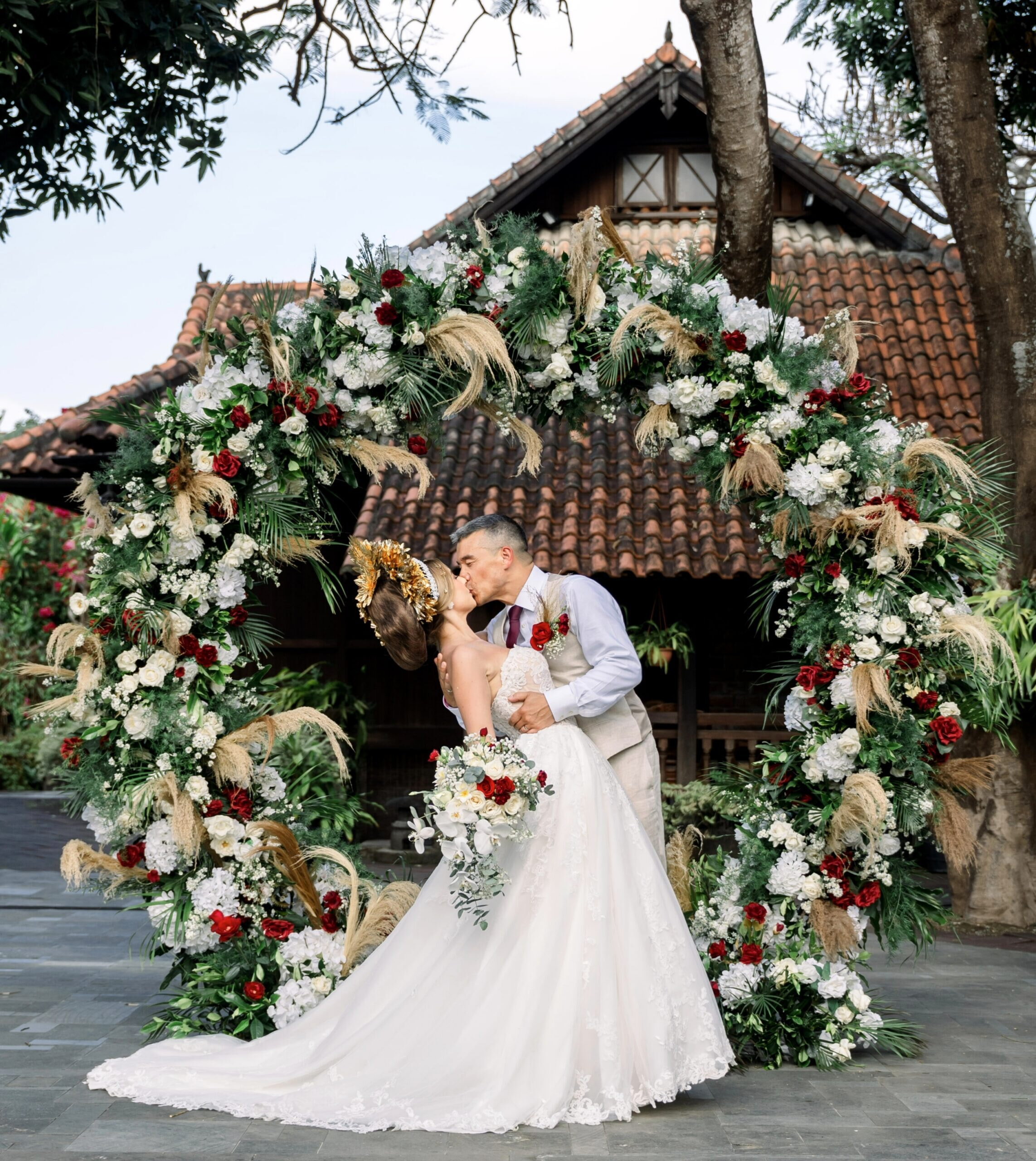 image of a bride and groom kissing infront of a florwar red and white rose wedding arch. the groom weaars a linnen suit and the bride wears a ballgown white dress with the bride is wearing Gelungan Payas Agung royal headress