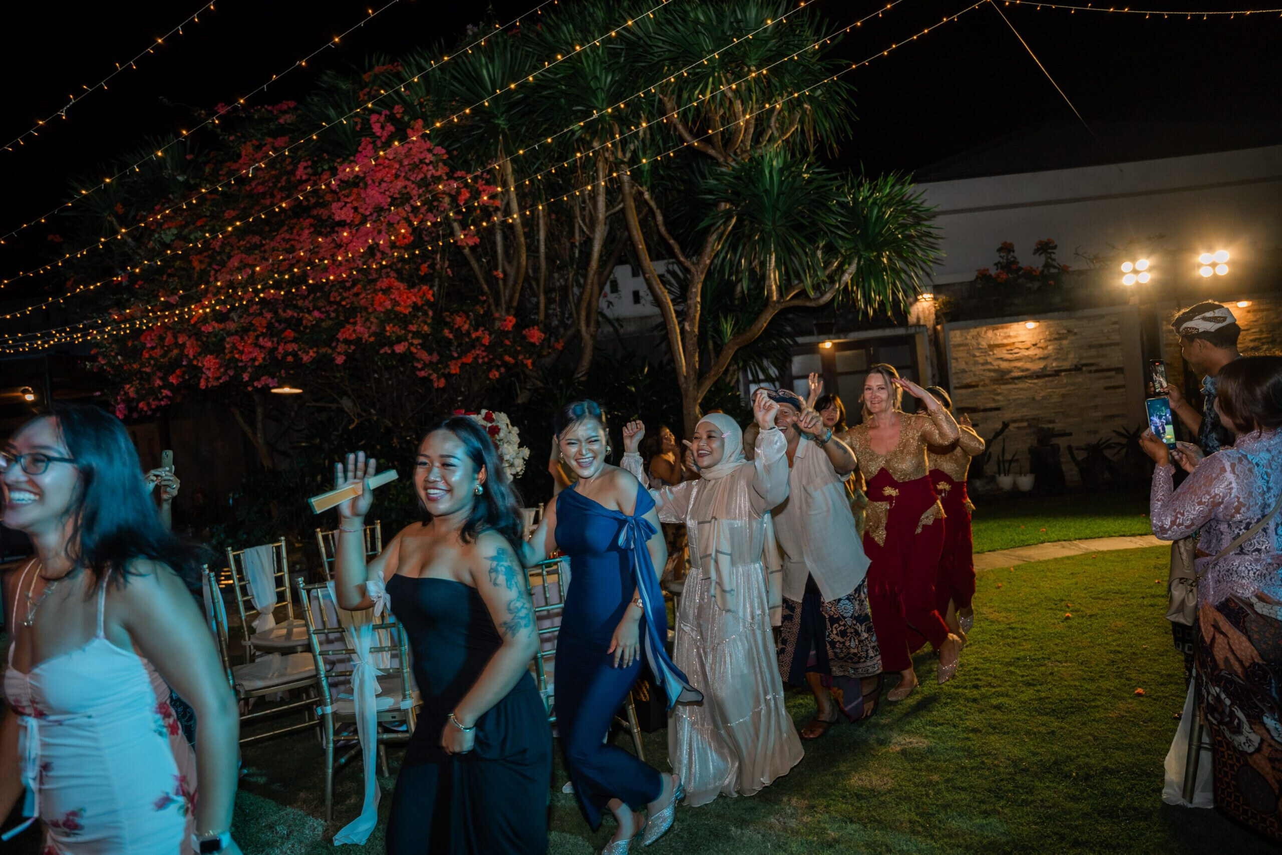 A line of ladies dressed in evening gowns dancing the conga at a night time wedding party
