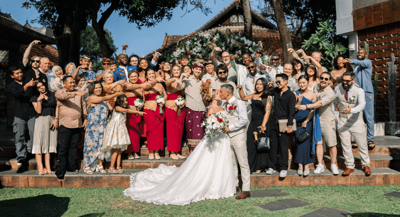 a photo of approximately a 45 person wedding congregation surrounding the bride and groom embracing kiss. The settingis tropical and a mixture of cultural attire is worn