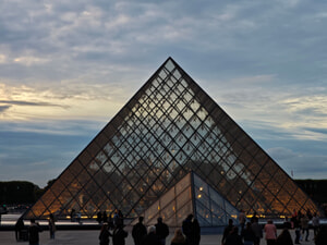 The glass pyramid entrance of the Louvre Museum in Paris at dusk, with visitors silhouetted in the foreground and a dramatic sky above.