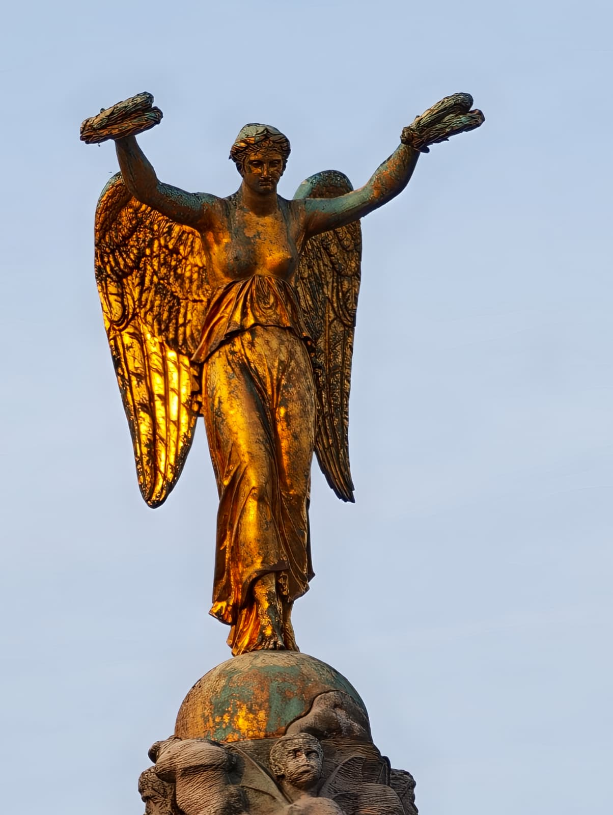 A golden angel statue holding two laurel wreaths stands atop a globe, glowing in the sunlight against a pale blue sky in Paris.