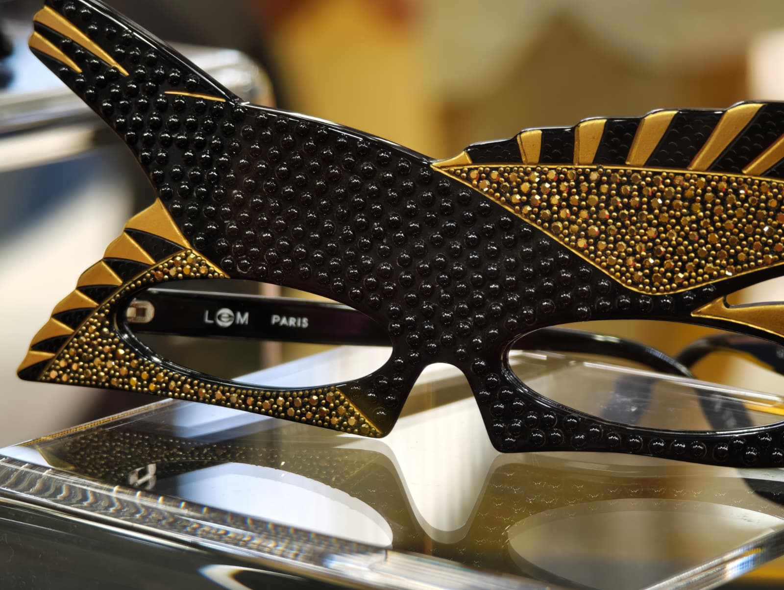 A dramatic, black and gold avant-garde eyewear front with geometric and studded details, displayed on a glass shelf at SILMO Paris 2025.